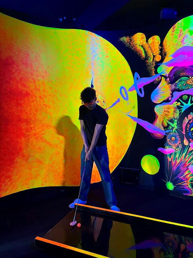 Eine Person spielt Mini-Golf in einem neonfarbenen, psychedelischen Raum mit leuchtenden Wänden, die von abstrakten Mustern und Farben dominiert werden. Die Person hält einen Golfschläger und zielt auf einen pinken Golfball.