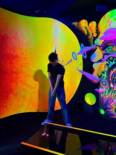 Eine Person spielt Mini-Golf in einem neonfarbenen, psychedelischen Raum mit leuchtenden Wänden, die von abstrakten Mustern und Farben dominiert werden. Die Person hält einen Golfschläger und zielt auf einen pinken Golfball.