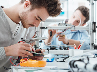 Zwei Personen arbeiten in einem Labor oder einer Werkstatt an elektronischen Komponenten. Die Person im Vordergrund hält eine Leiterplatte und nutzt eine Lupe, während die Person im Hintergrund offenbar mit einem 3D-Drucker oder einem ähnlichen Gerät arbeitet. Auf dem Tisch befinden sich verschiedene Werkzeuge und elektronische Geräte, darunter ein orangefarbenes Multimeter, Kabel sowie Behälter mit Stiften und anderen Kleinteilen.